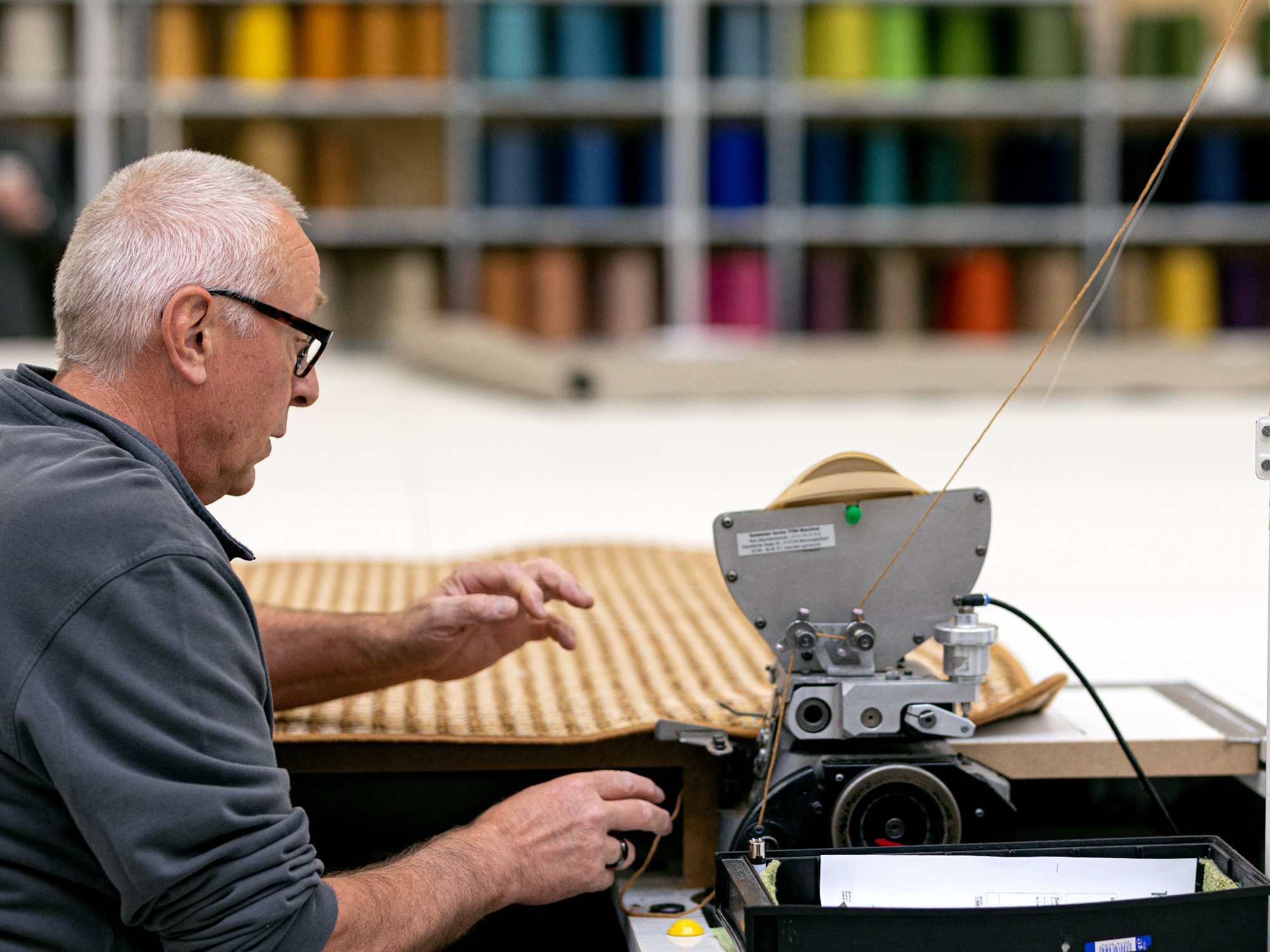 Mitarbeiter näht Teppichkante an einer Nähmaschine, im Hintergrund bunte Bordürenrollen