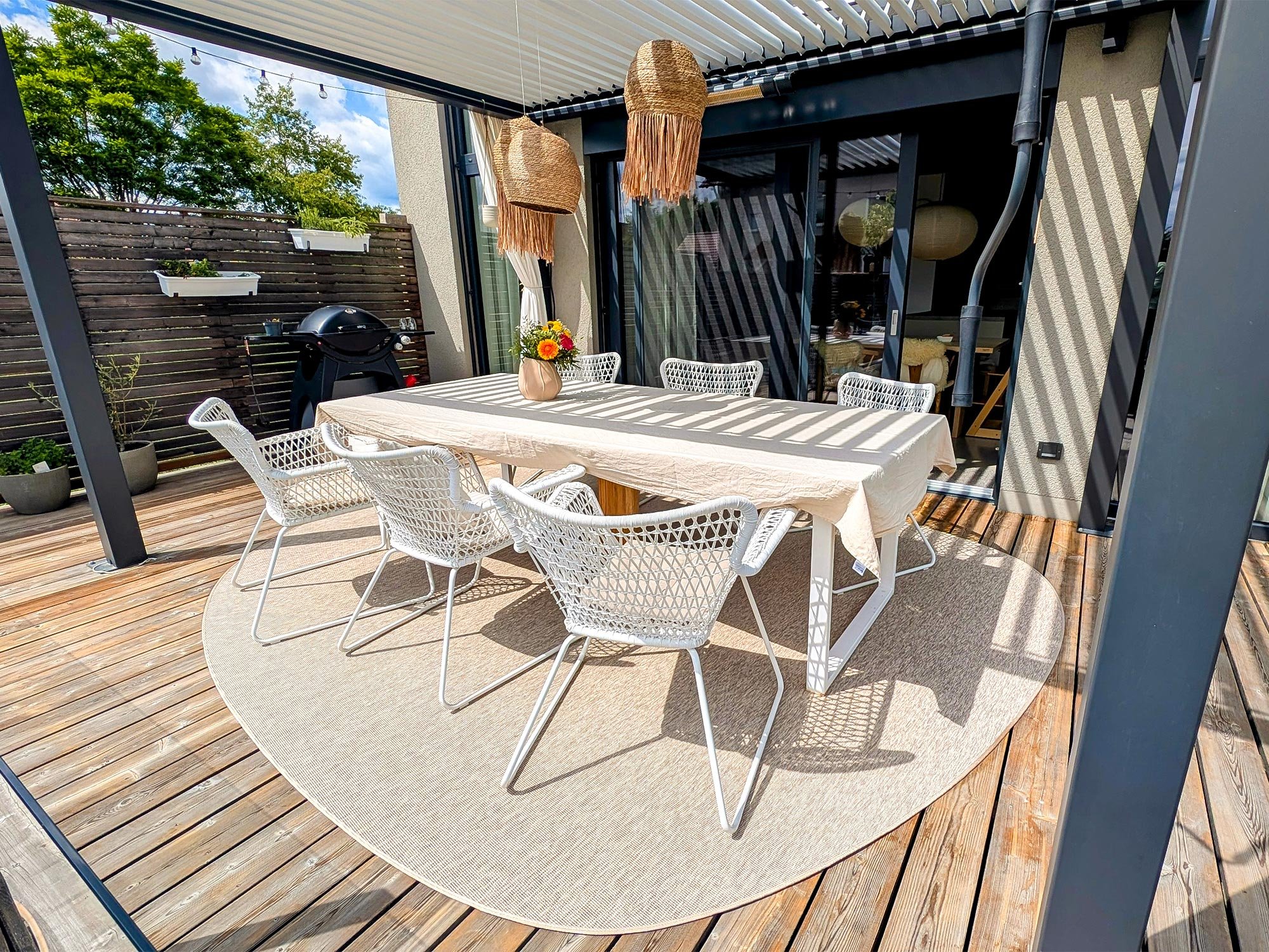 Beiger Pro Nature Outdoorteppich in organischer Form auf moderner Holzterrasse mit Esstisch und weißen Stühlen unter Pergola