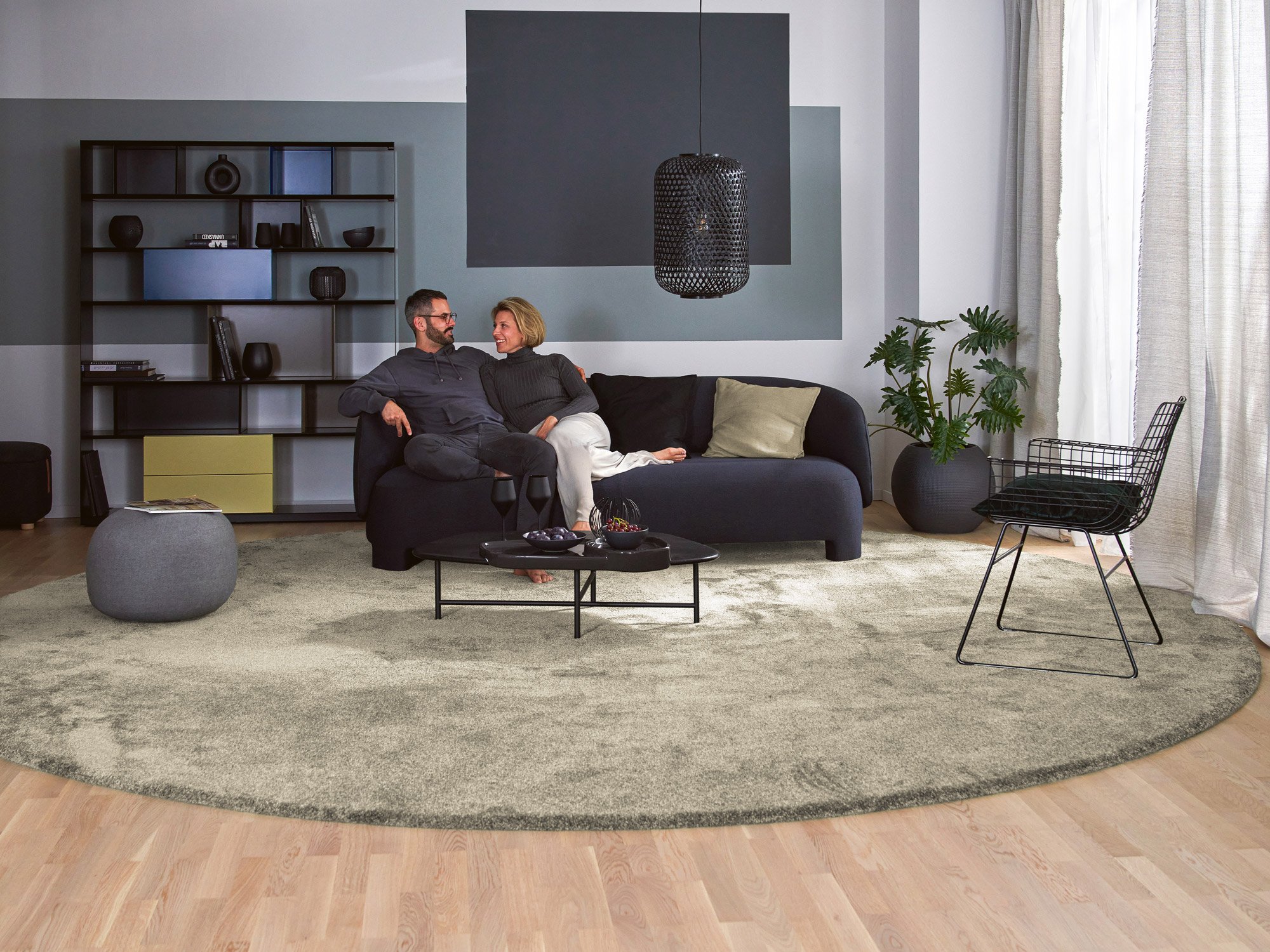 Modernes Wohnzimmer mit rundem Hochflor-Coverteppich Brienne in Greige von Teppichscheune, vor dunklem Sofa und dekorativen Akzenten