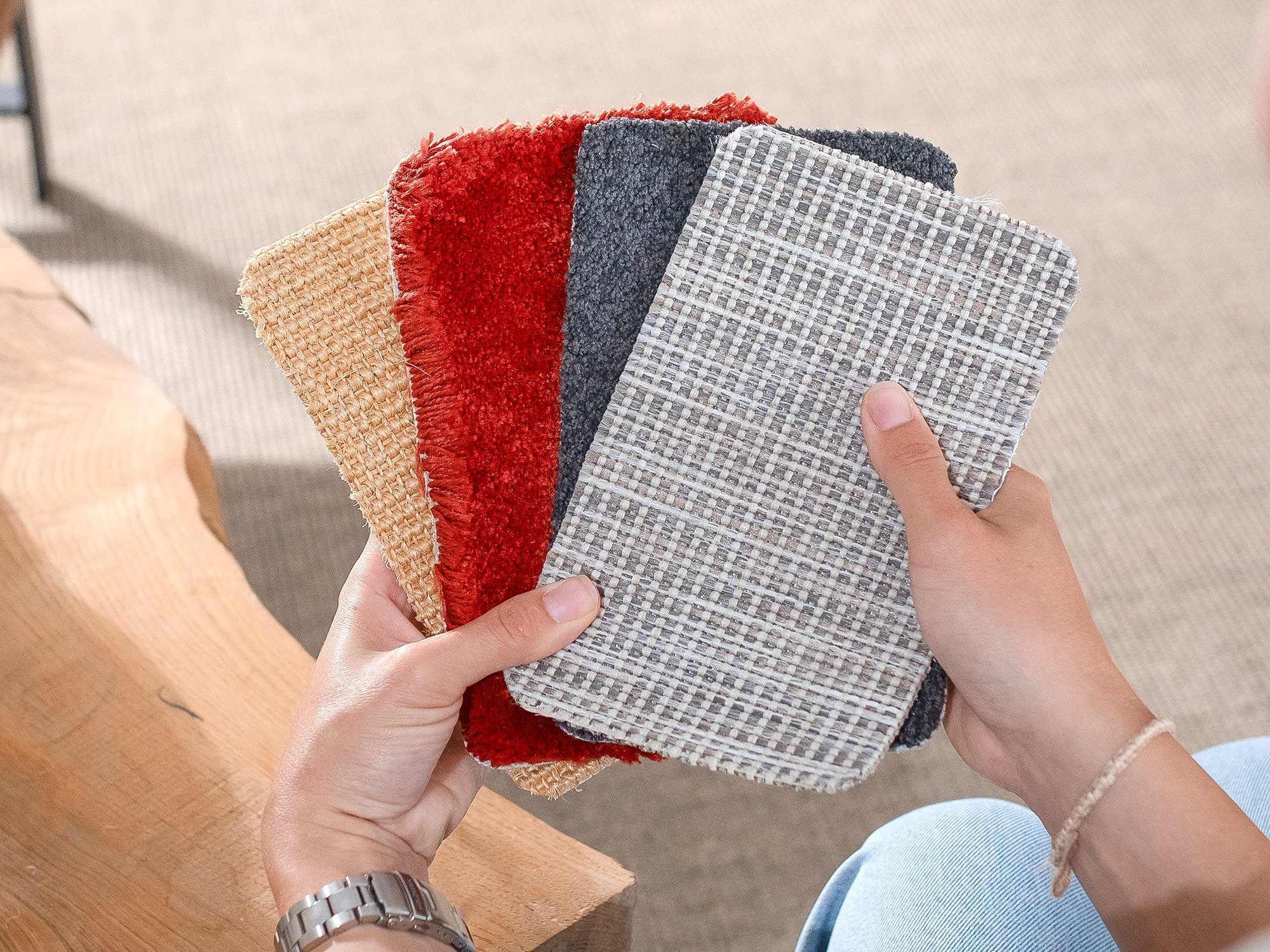 Eine Hand hält vier Teppichmuster unterschiedlicher Materialien (Sisal, Hochflor, Kurzflor und Outdoor) in die Kamera